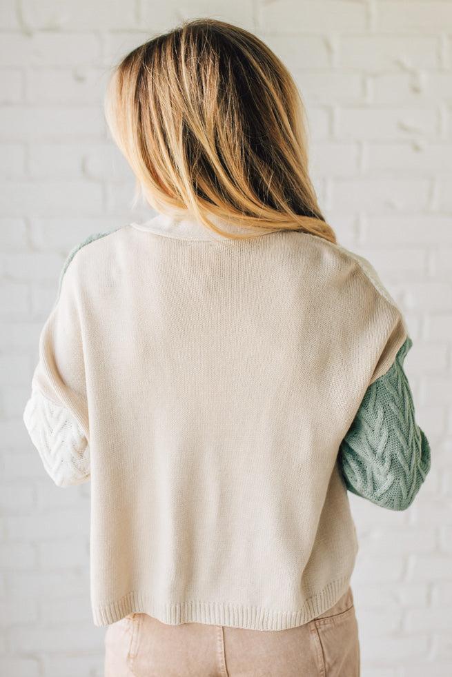 Person wearing a beige sweater vest over a green shirt against a white brick wall.