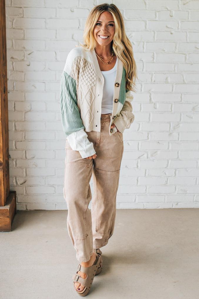 Woman wearing a light-colored jacket, white top, and beige pants against a white brick wall.
