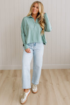 Woman wearing a light green shirt and light blue jeans standing against a white paneled wall.