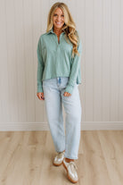 Woman wearing a green long-sleeve shirt and light blue jeans standing against a white paneled wall.