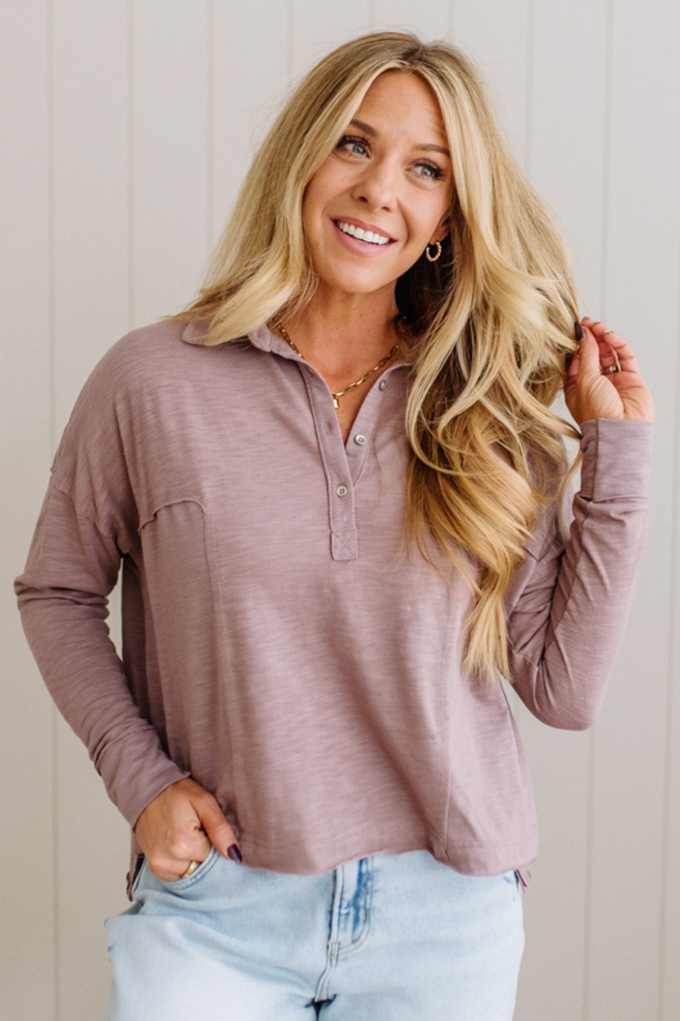 Woman wearing a mauve long-sleeve shirt and light blue jeans against a white wall.