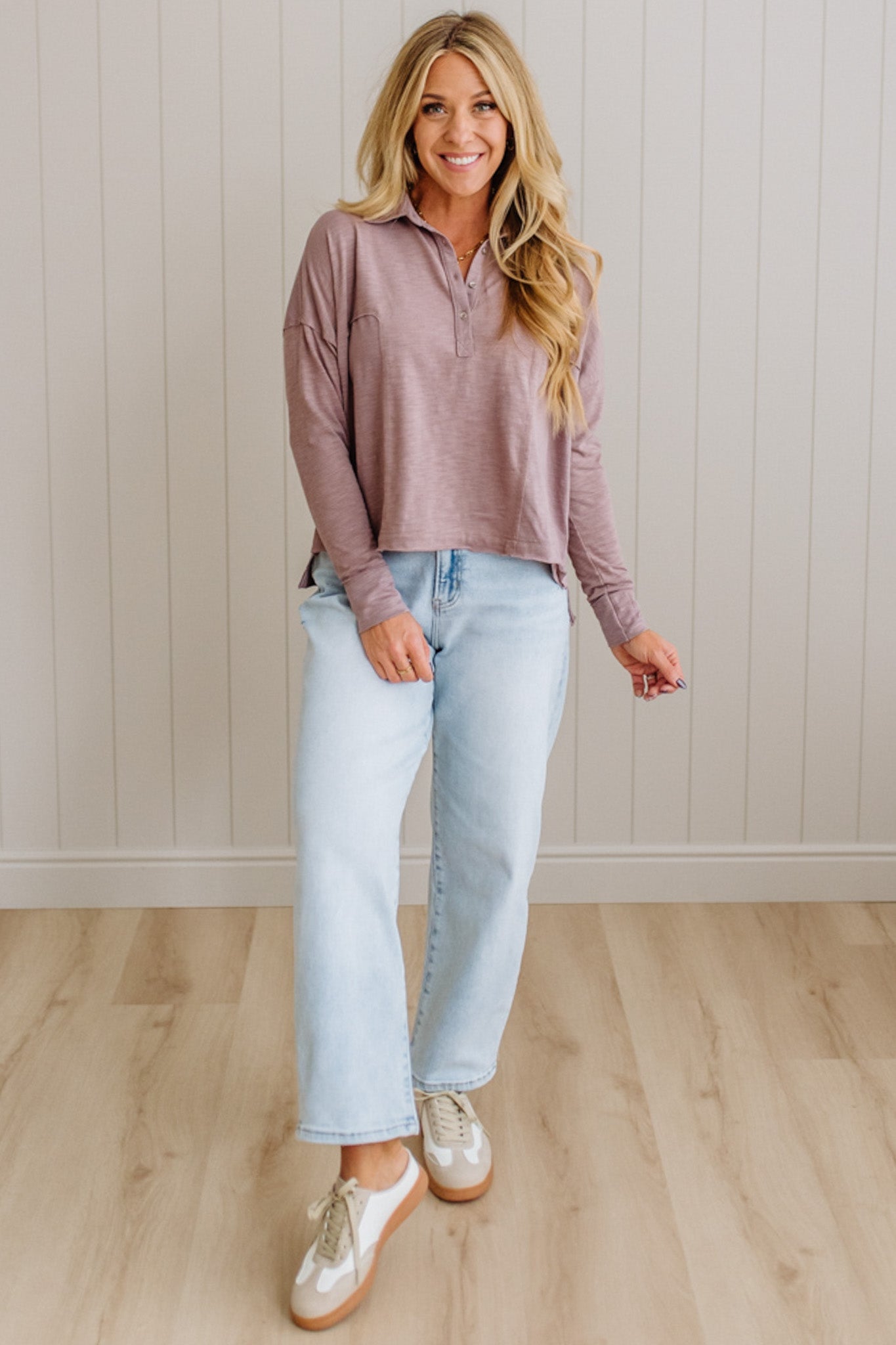 Woman wearing a mauve long-sleeve top and light blue jeans standing against a white paneled wall.