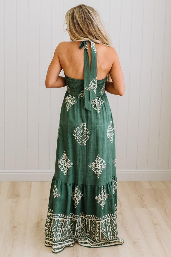 Woman wearing a green dress with white patterns in a room with wooden flooring and a white wall.