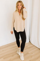 Woman wearing a beige long-sleeve top, black leggings, and white sneakers in a minimalistic room.