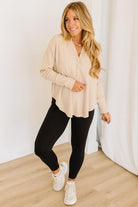 Woman wearing a beige top and black leggings standing against a white curtain.