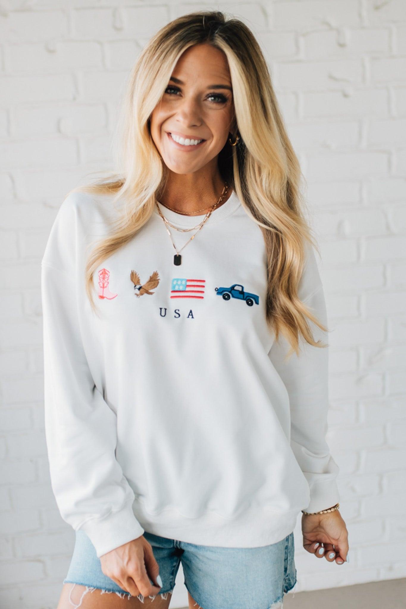 Blonde woman in denim shorts wearing a white pullover with embroidery details  of a boot, eagle, flag and truck with USA under them.