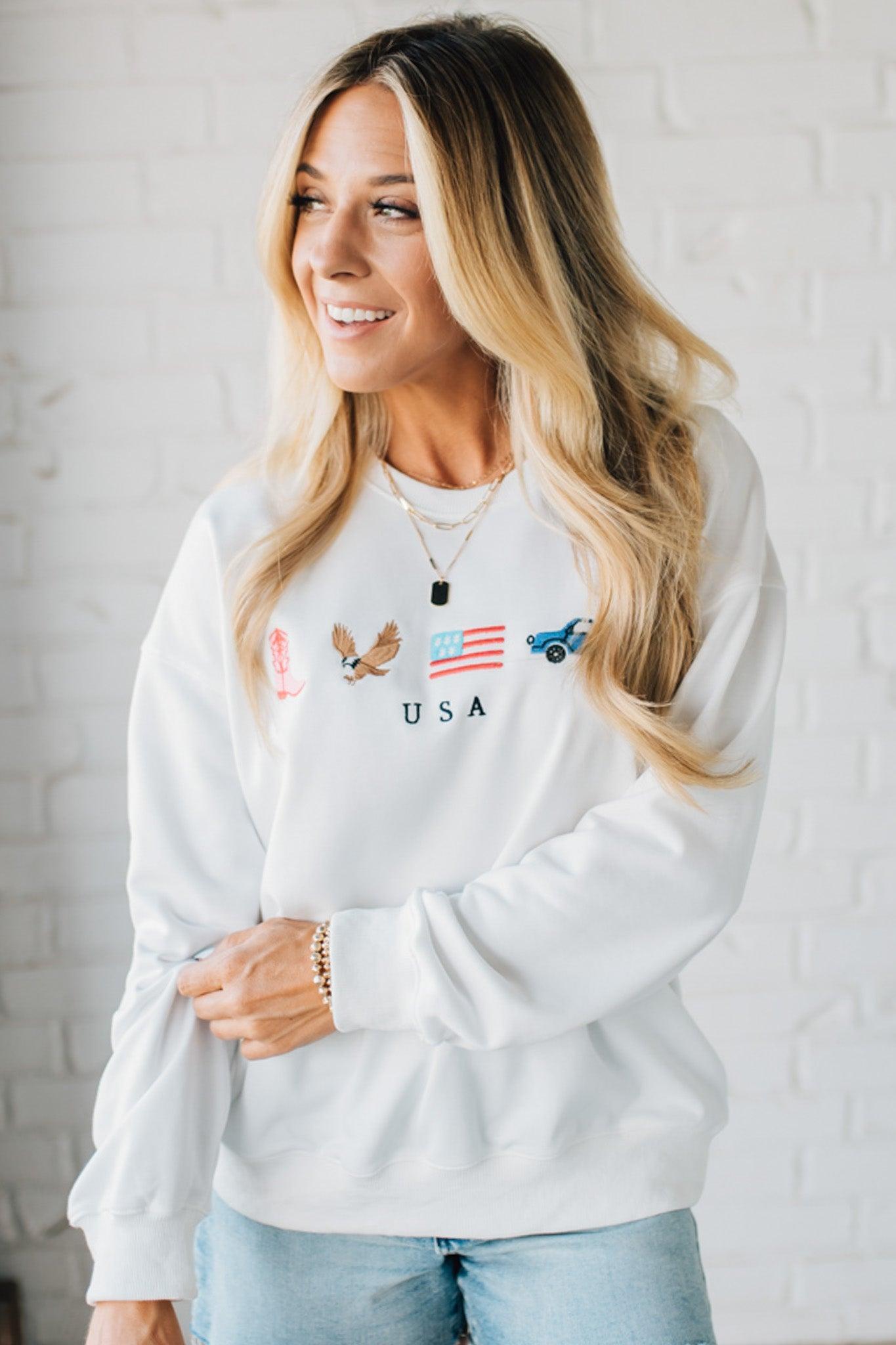 Blonde woman in denim shorts wearing a white pullover with embroidery details  of a boot, eagle, flag and truck with USA under them.