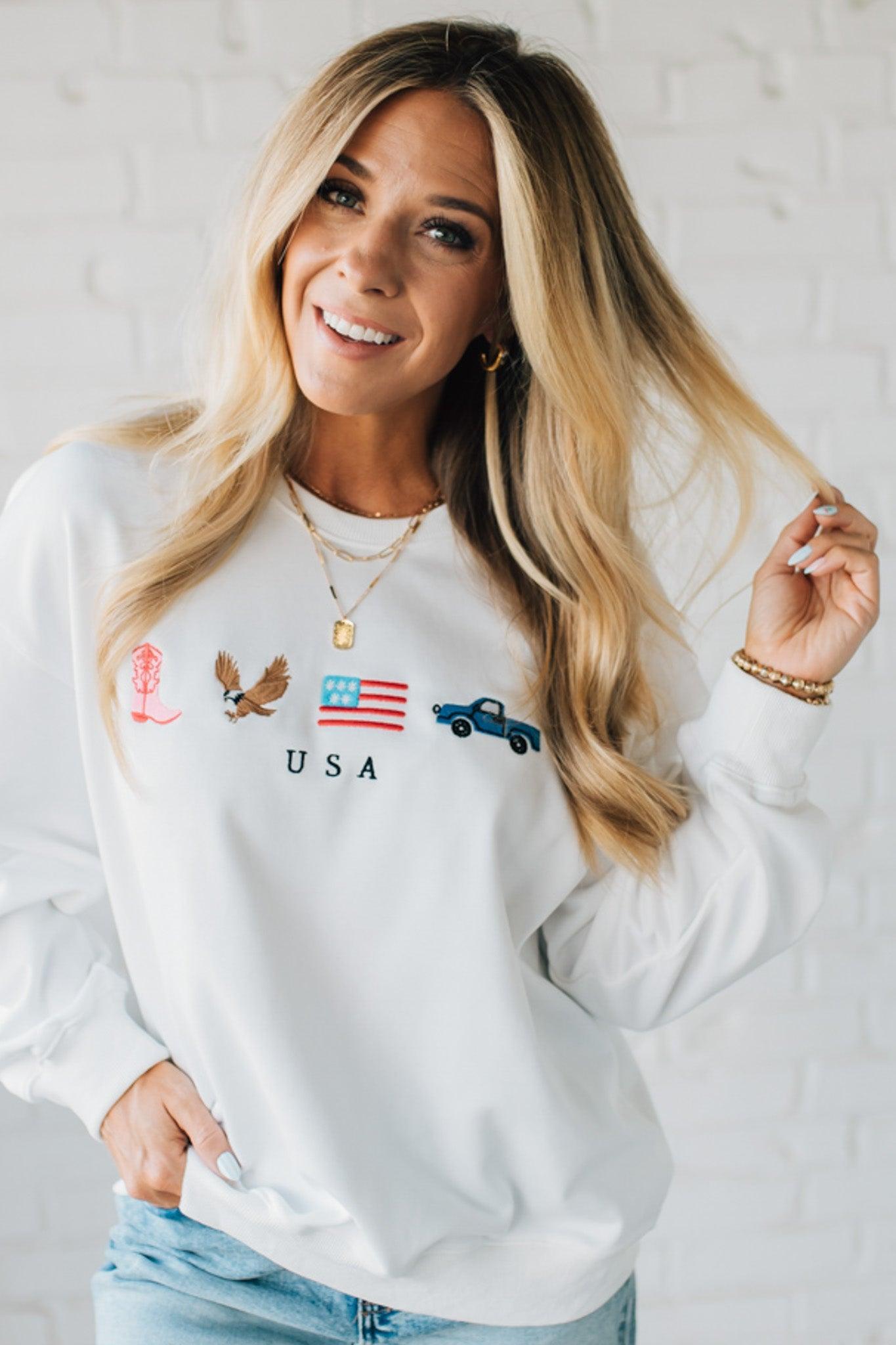 Blonde woman in denim shorts wearing a white pullover with embroidery details  of a boot, eagle, flag and truck with USA under them.