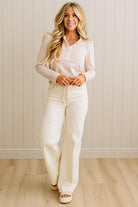 Woman wearing a striped shirt and white pants standing against a light wooden paneled wall.
