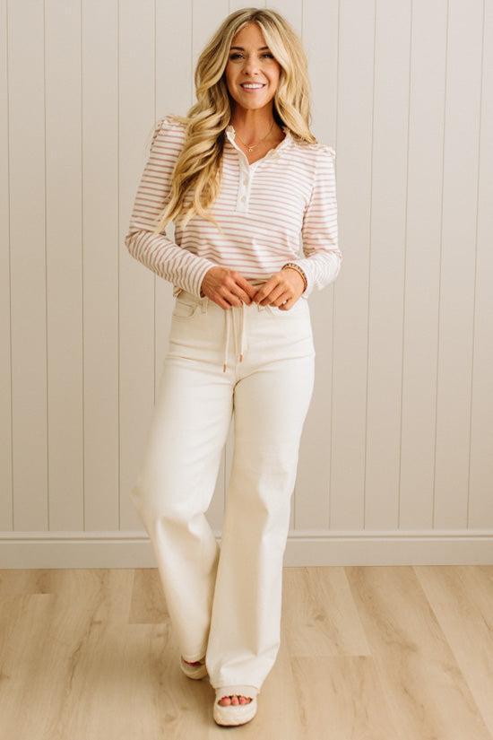 Woman wearing a striped shirt and white pants standing against a light wooden paneled wall.