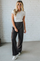 Woman wearing a white t-shirt and dark pants standing against a white brick wall.