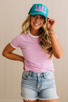 Woman wearing a pink striped shirt, denim shorts, and a turquoise cap with 'LAKE' on it against a beige background.