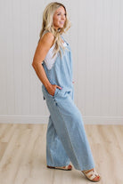 Woman wearing light blue denim overalls standing against a white paneled wall.