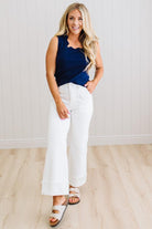 Woman wearing a navy top and white pants standing against a light wooden floor and white wall.