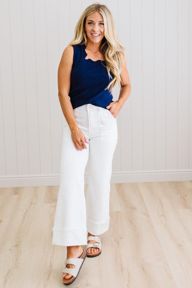 Woman wearing a navy top and white pants standing against a light wooden floor and white wall.
