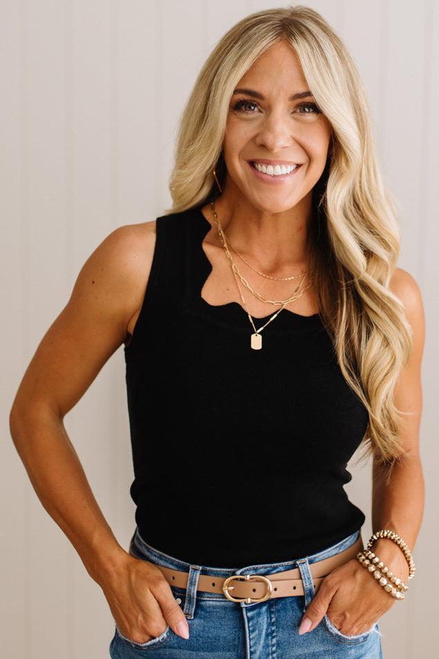 A smiling blonde woman wearing a black sleeveless scoop-neck top, blue jeans, and gold necklaces, standing against a light beige wall.