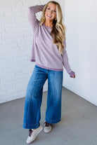 Blonde woman against a white wall wearing a lavender and white striped sweater with blue jeans and sneakers.