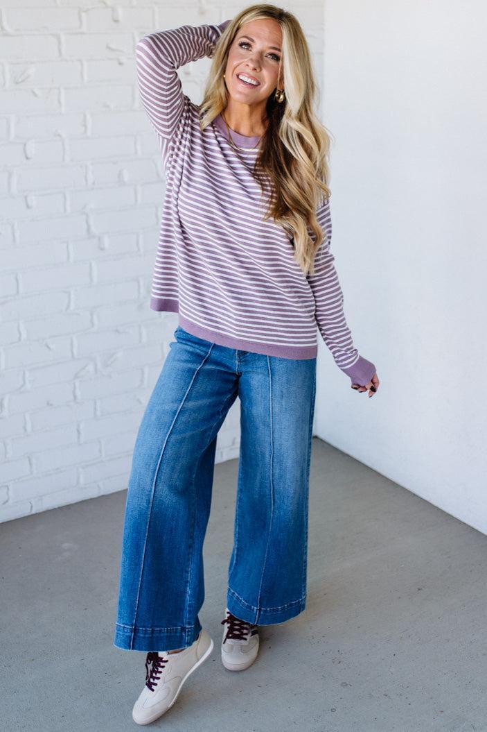 Blonde woman against a white wall wearing a lavender and white striped sweater with blue jeans and sneakers.