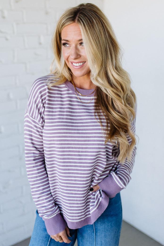 Blonde woman against a white wall wearing a lavender and white striped sweater with blue jeans and sneakers.