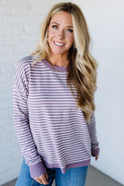Blonde woman against a white wall wearing a lavender and white striped sweater with blue jeans and sneakers.