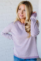 Blonde woman against a white wall wearing a lavender and white striped sweater with blue jeans and sneakers.