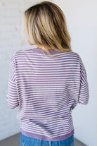 Blonde woman against a white wall wearing a lavender and white striped sweater with blue jeans and sneakers.
