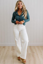 Woman wearing a teal top and white pants against a light wooden paneled wall.