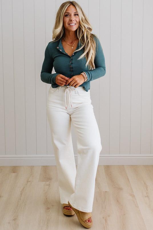 Woman wearing a teal top and white pants against a light wooden paneled wall.