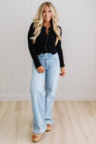 Woman wearing a black long-sleeve top and light blue jeans standing against a white paneled wall.