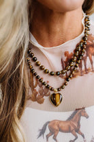 A close-up of a woman wearing a double-strand beaded necklace with brown and black tiger's eye beads and a heart-shaped pendant, paired with a white t-shirt featuring a brown horse print.