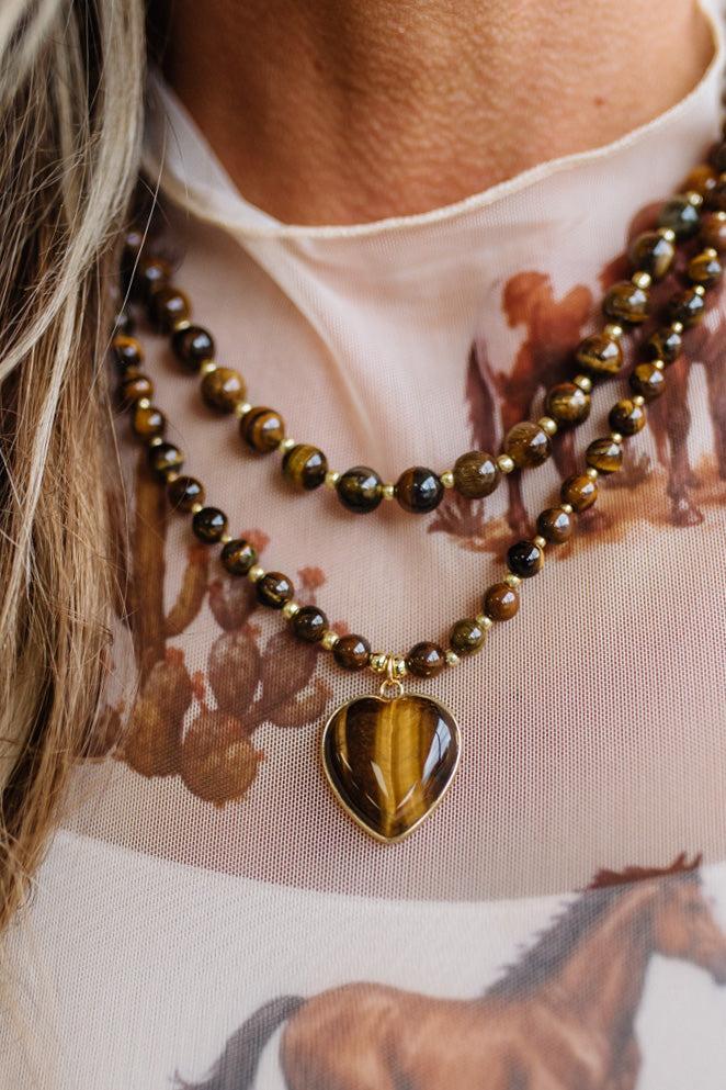 A close-up of a person wearing a double-strand beaded necklace with a heart-shaped tiger's eye pendant, layered over a sheer printed top.