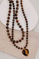 A double-strand beaded necklace featuring tiger's eye beads in shades of brown, black, and gold, with a heart-shaped tiger's eye pendant in a gold-tone bezel, displayed on a textured light beige surface.