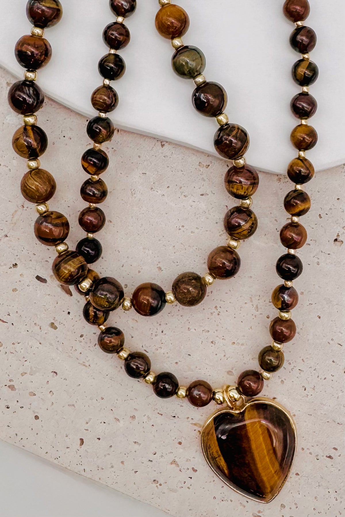 A double-strand beaded necklace featuring tiger's eye beads in shades of brown, black, and gold, with a heart-shaped tiger's eye pendant in a gold-tone bezel, displayed on a textured light beige surface.