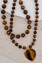 A double-strand beaded necklace featuring tiger's eye beads in shades of brown, black, and gold, with a heart-shaped tiger's eye pendant in a gold-tone bezel, displayed on a textured light beige surface.