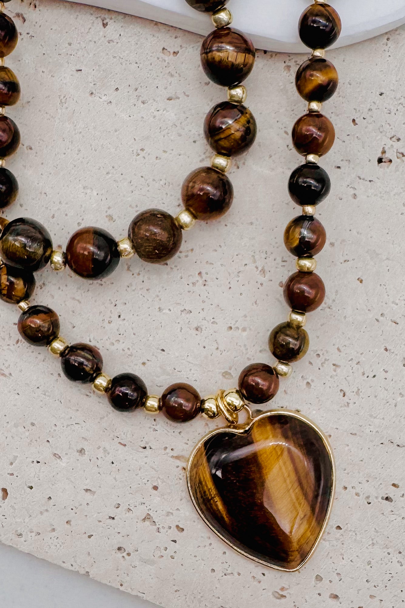 A double-strand beaded necklace featuring tiger's eye beads in shades of brown, black, and gold, with a heart-shaped tiger's eye pendant in a gold-tone bezel, displayed on a textured light beige surface.
