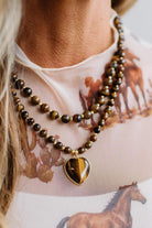 A close-up of a person wearing a double-strand beaded necklace with polished brown and black tiger's eye beads and a heart-shaped pendant, paired with a light beige printed top.