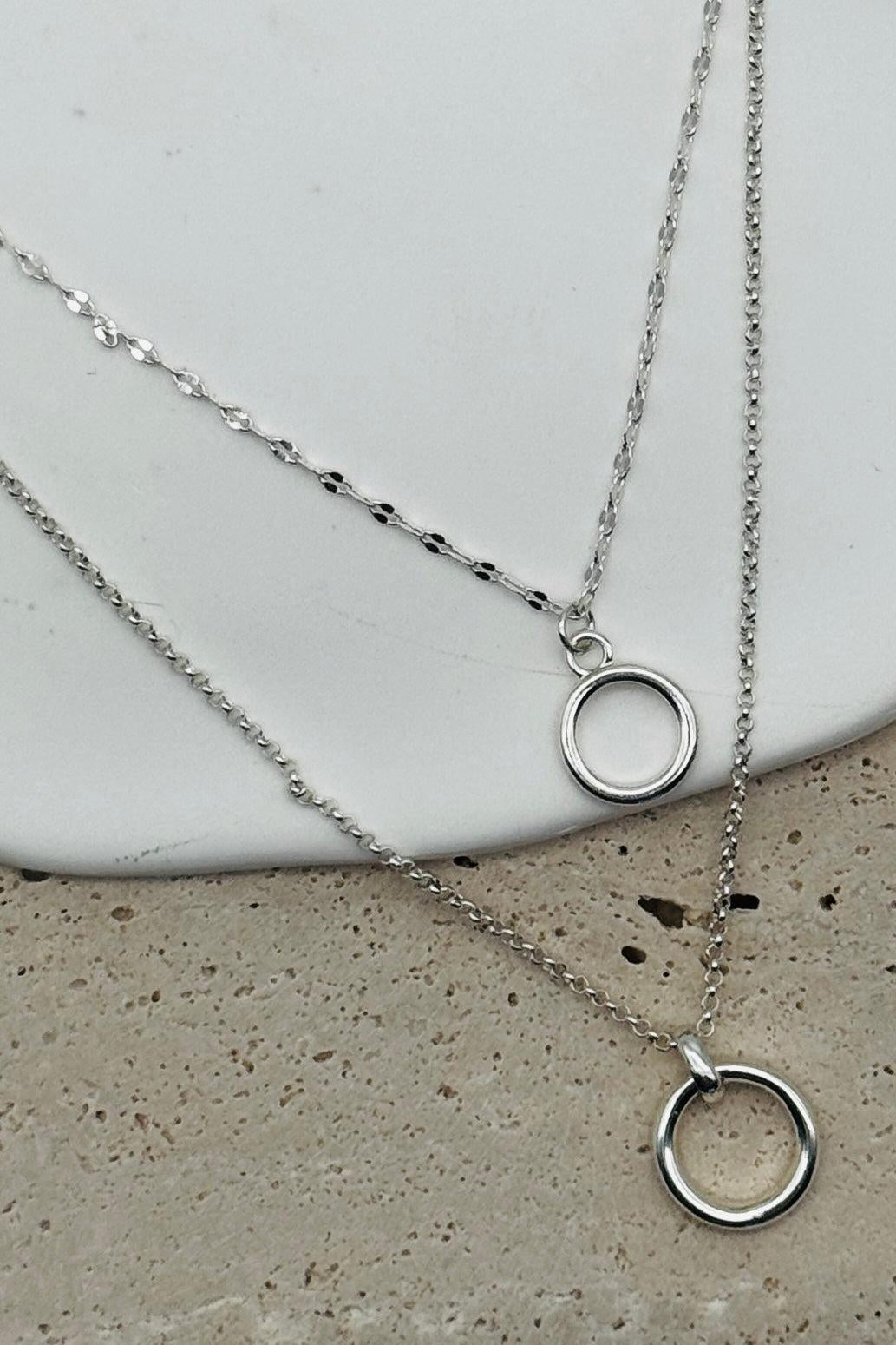 Two silver necklaces with circular pendants on a concrete surface.