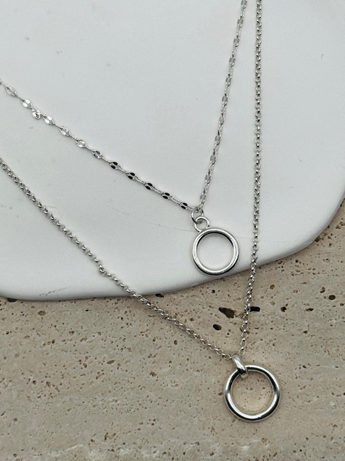 Two silver necklaces with circular pendants on a concrete surface.
