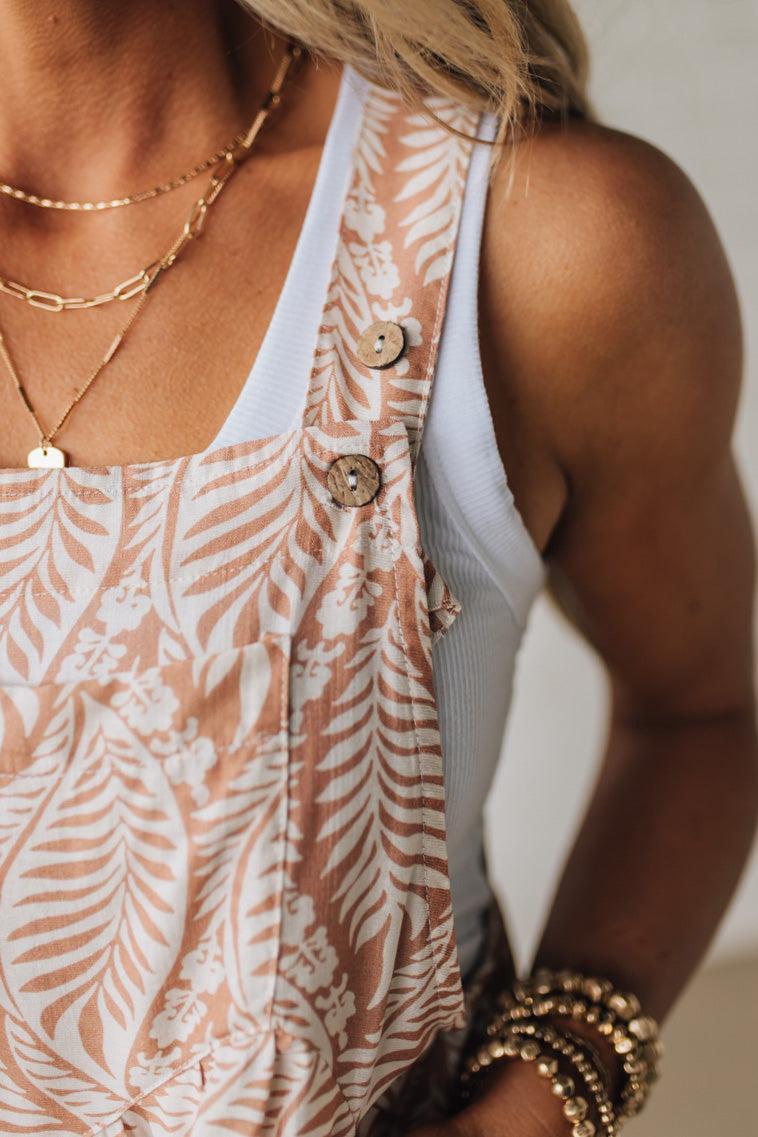 Blonde woman wearing short overalls with wide leg, triple button adjustable straps, and all over leafy print in coral and white.