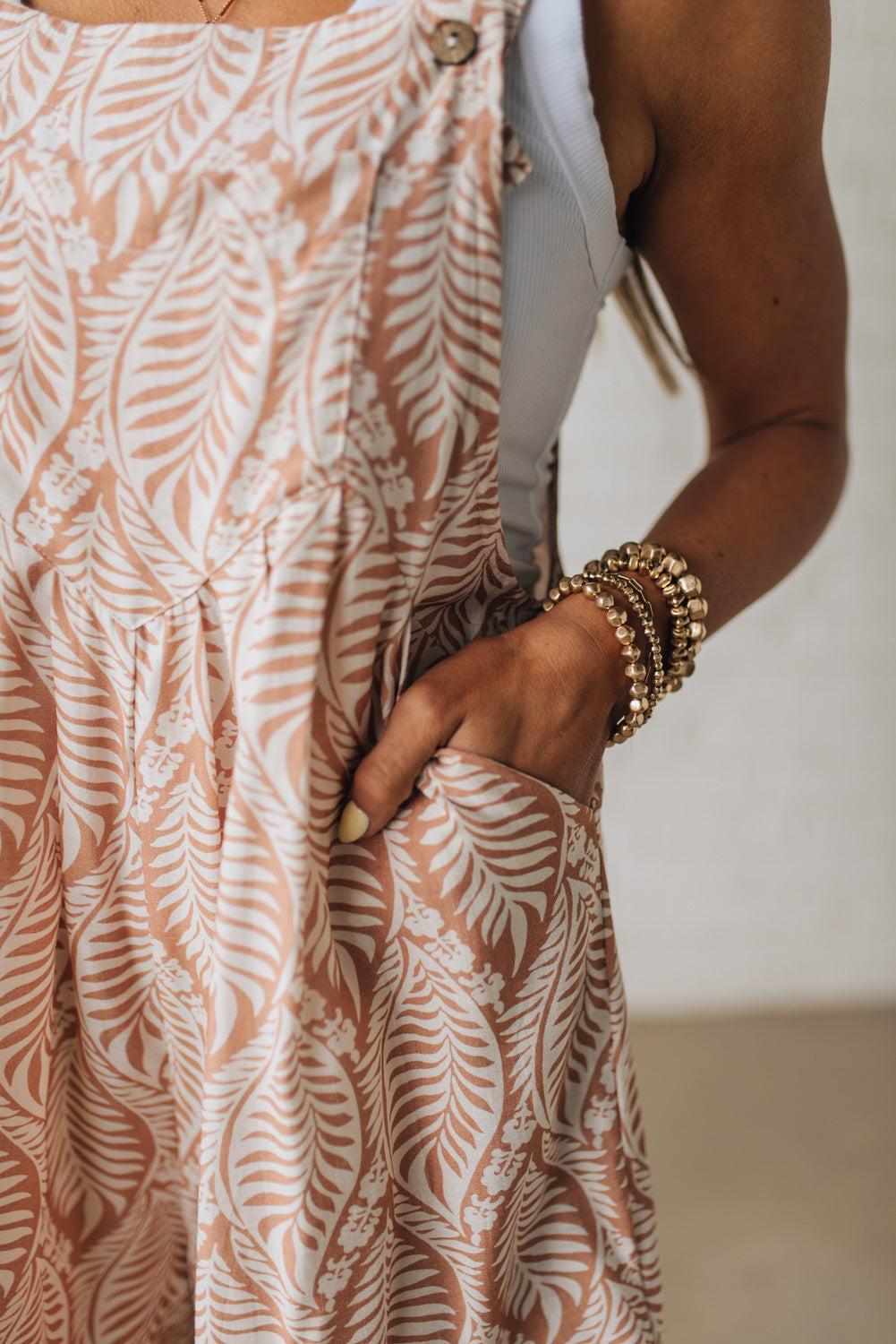 Blonde woman wearing short overalls with wide leg, triple button adjustable straps, and all over leafy print in coral and white.