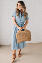 Blonde woman in blue dress holding an oversized rectangle straw tote with wood handles.