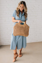 Blonde woman in blue dress holding an oversized rectangle straw tote with wood handles.