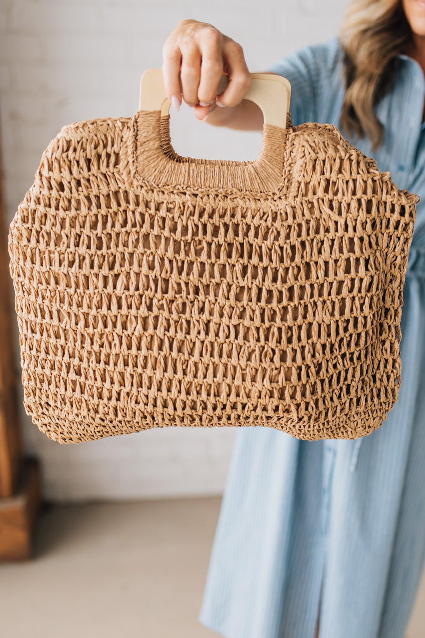 Blonde woman in blue dress holding an oversized rectangle straw tote with wood handles.