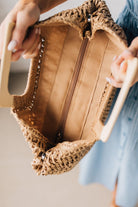 Blonde woman in blue dress holding an oversized rectangle straw tote with wood handles.