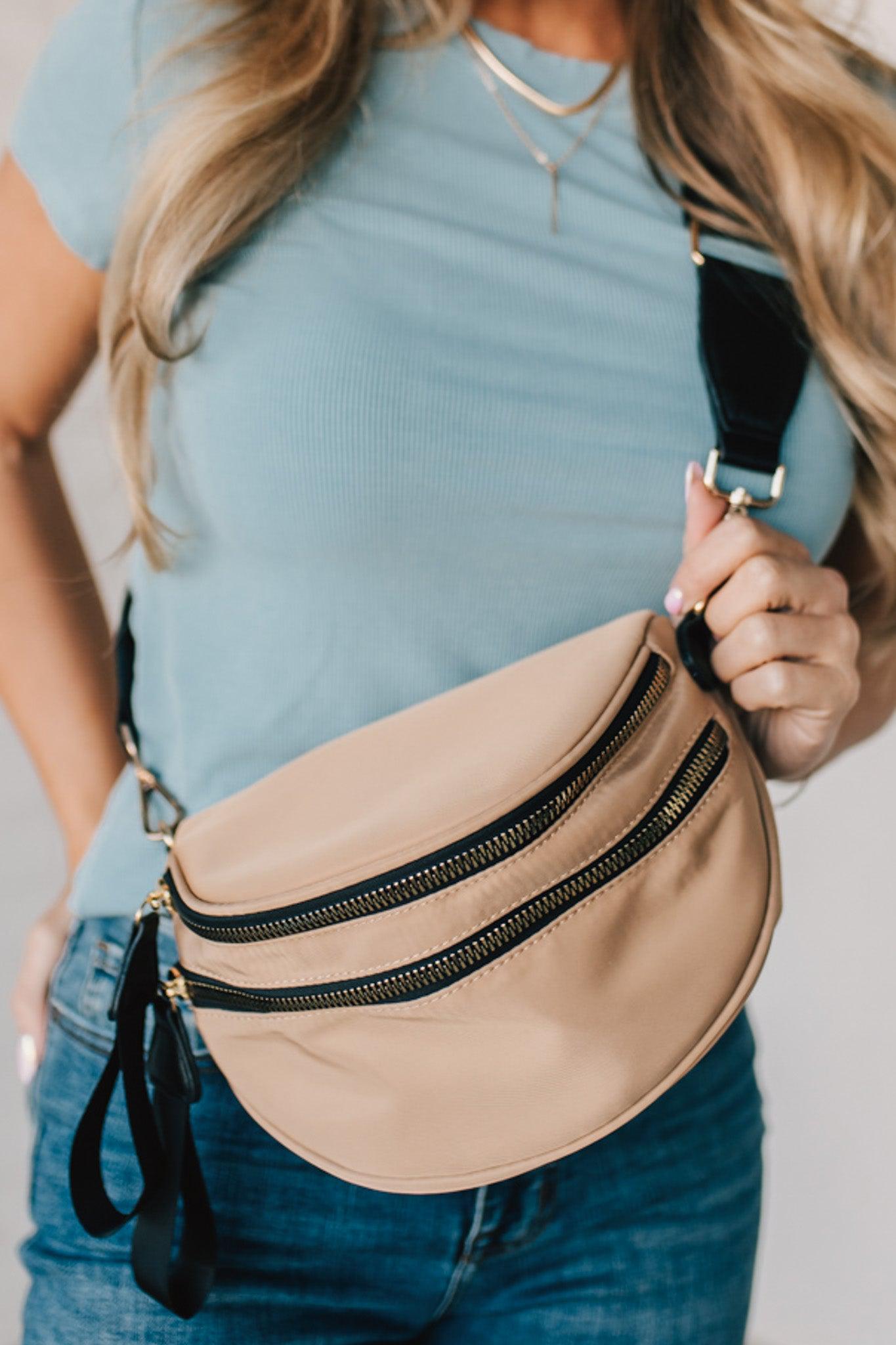 Blonde woman in blue shirt wearing a tan crossbody bag with black zippers.