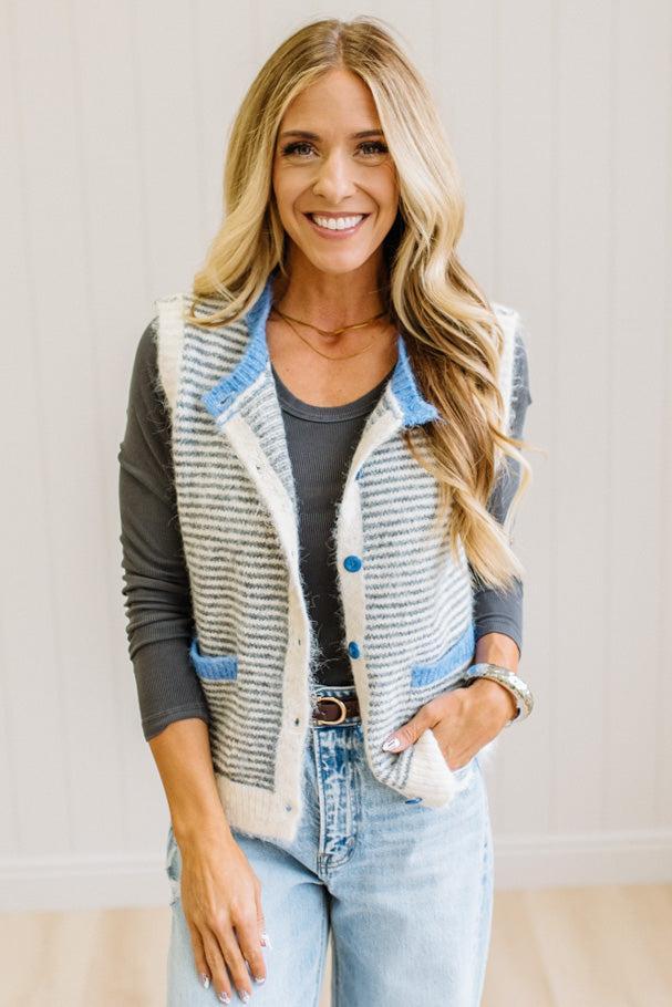 BLONDE WOMAN IN LIGHT JEANS WEARING A DARK GREY TOP WITH A FUZZY SWEATER VEST THAT HAS BLUE TRIM CONTRAST AND BUTTONS, AND A CREAM AND GREY STRIPED BODY.