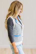 BLONDE WOMAN IN LIGHT JEANS WEARING A DARK GREY TOP WITH A FUZZY SWEATER VEST THAT HAS BLUE TRIM CONTRAST AND BUTTONS, AND A CREAM AND GREY STRIPED BODY.