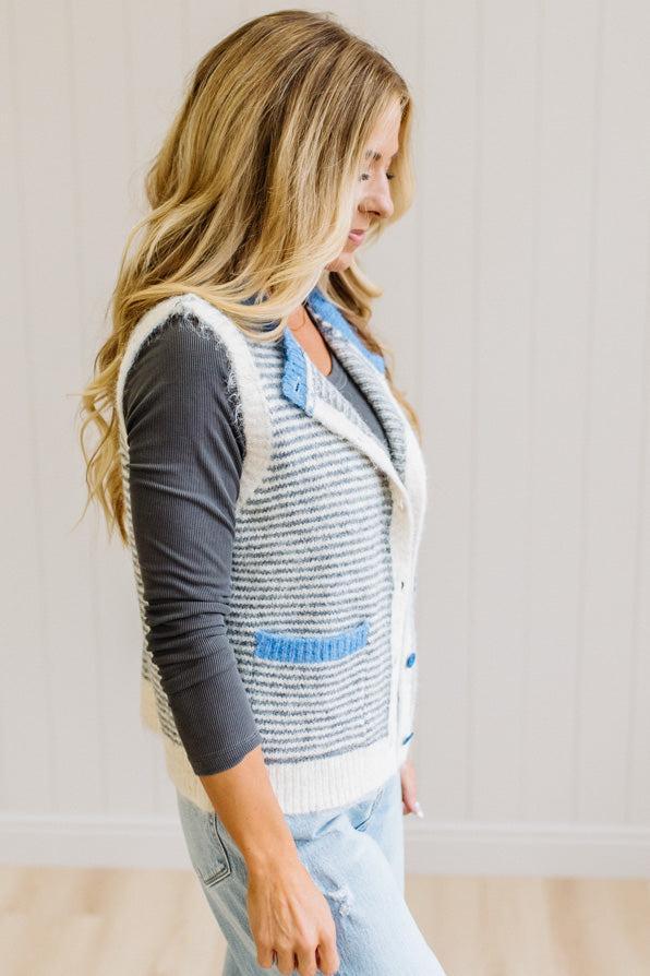 BLONDE WOMAN IN LIGHT JEANS WEARING A DARK GREY TOP WITH A FUZZY SWEATER VEST THAT HAS BLUE TRIM CONTRAST AND BUTTONS, AND A CREAM AND GREY STRIPED BODY.