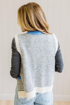BLONDE WOMAN IN LIGHT JEANS WEARING A DARK GREY TOP WITH A FUZZY SWEATER VEST THAT HAS BLUE TRIM CONTRAST AND BUTTONS, AND A CREAM AND GREY STRIPED BODY.
