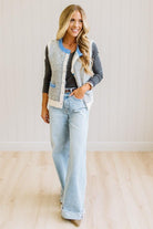BLONDE WOMAN IN LIGHT JEANS WEARING A DARK GREY TOP WITH A FUZZY SWEATER VEST THAT HAS BLUE TRIM CONTRAST AND BUTTONS, AND A CREAM AND GREY STRIPED BODY.